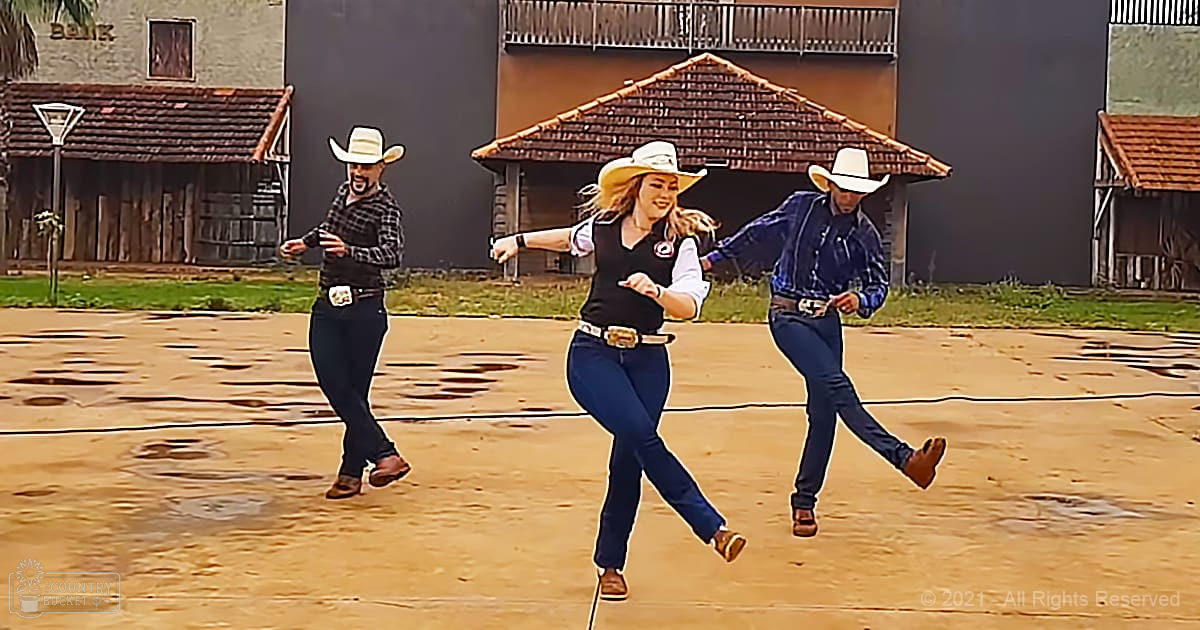 Group dancing outdoors in a rural small-town setting with buildings, a bank sign, lampposts, and grass under soft late afternoon or early evening light.