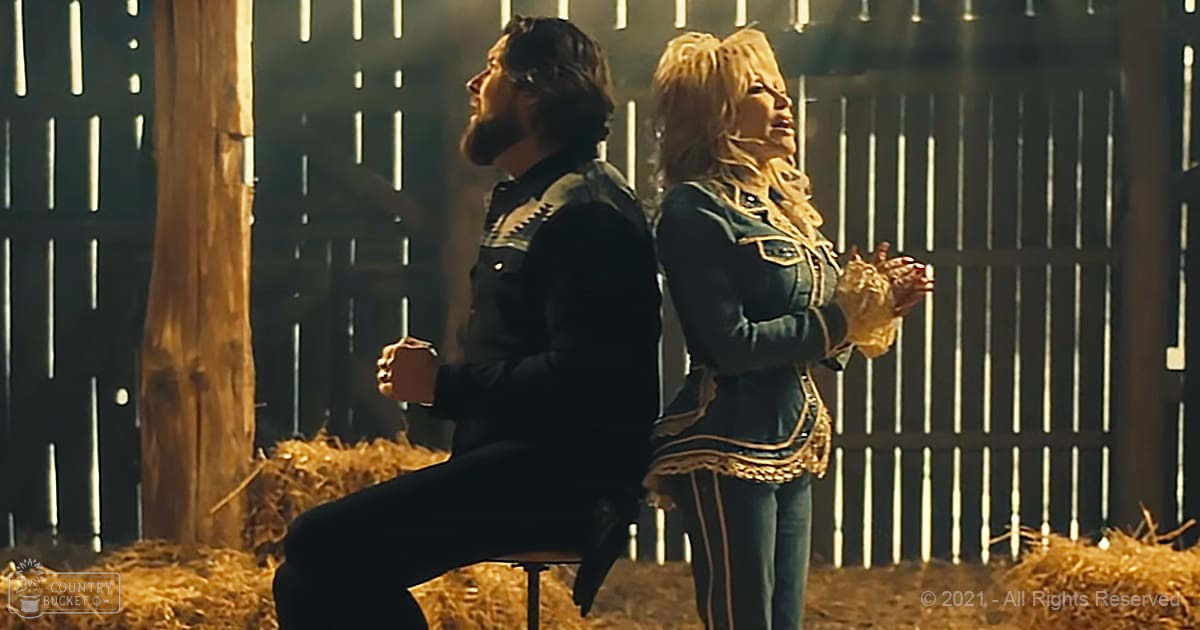 Two adults, a man and a woman, sit on hay bales inside a barn with wooden slats; the man wears black pants and a denim shirt with white accents, while the woman sports a blue jacket with gold trim and jeans with thin stripes down the sides.