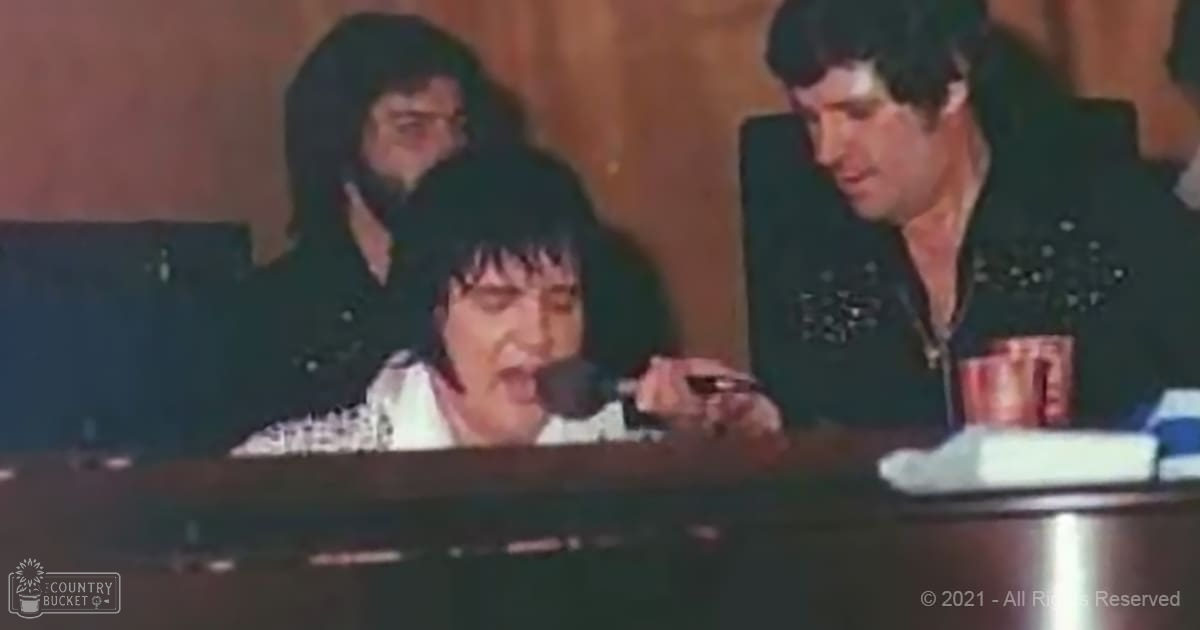 A woman singing into a microphone, accompanied by two men in similar jackets, set against an indoor wooden panel wall with artificial lighting.