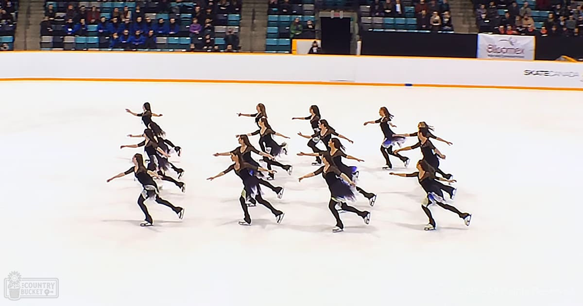 Synchronized skating team performing at an indoor ice rink with young adult female skaters wearing black dresses with white and blue accents, seated spectators, well-lit artificial lighting.