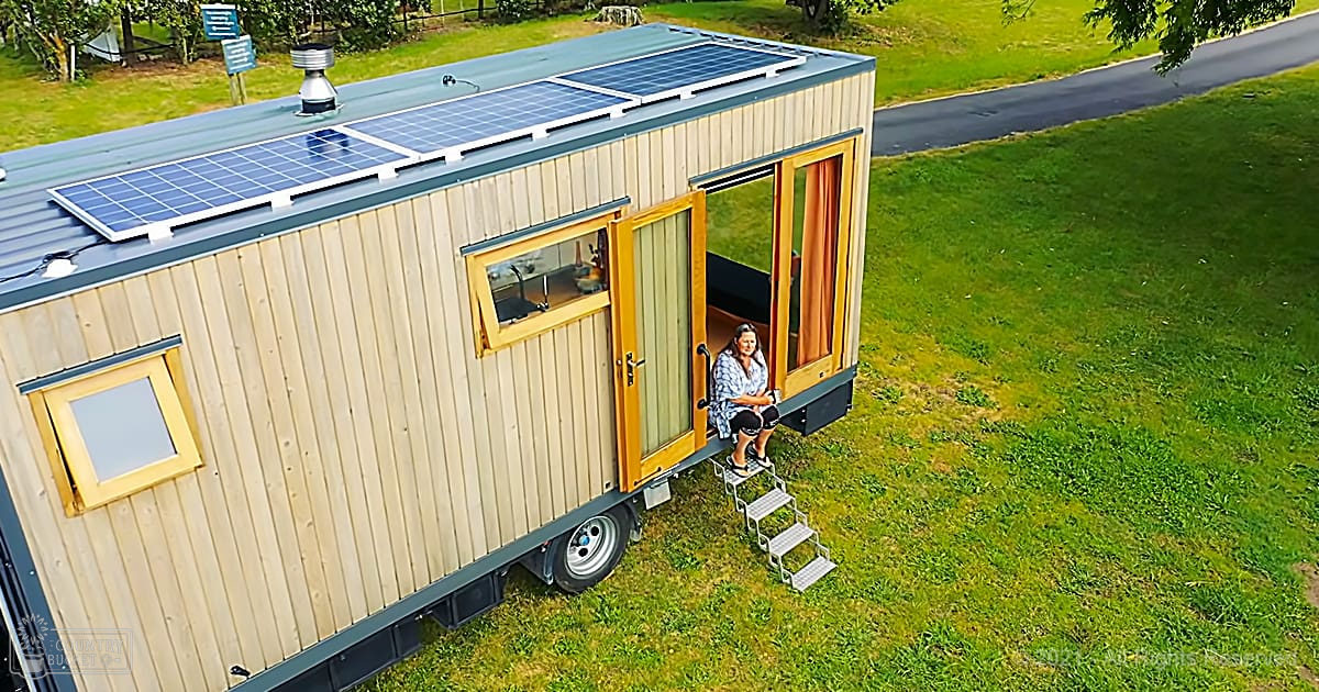 A woman in a casual blue floral shirt and shorts sits on a small step ladder leading to the door of a tiny house with solar panels. The scene is outdoors, likely spring or summer, with bright daylight.