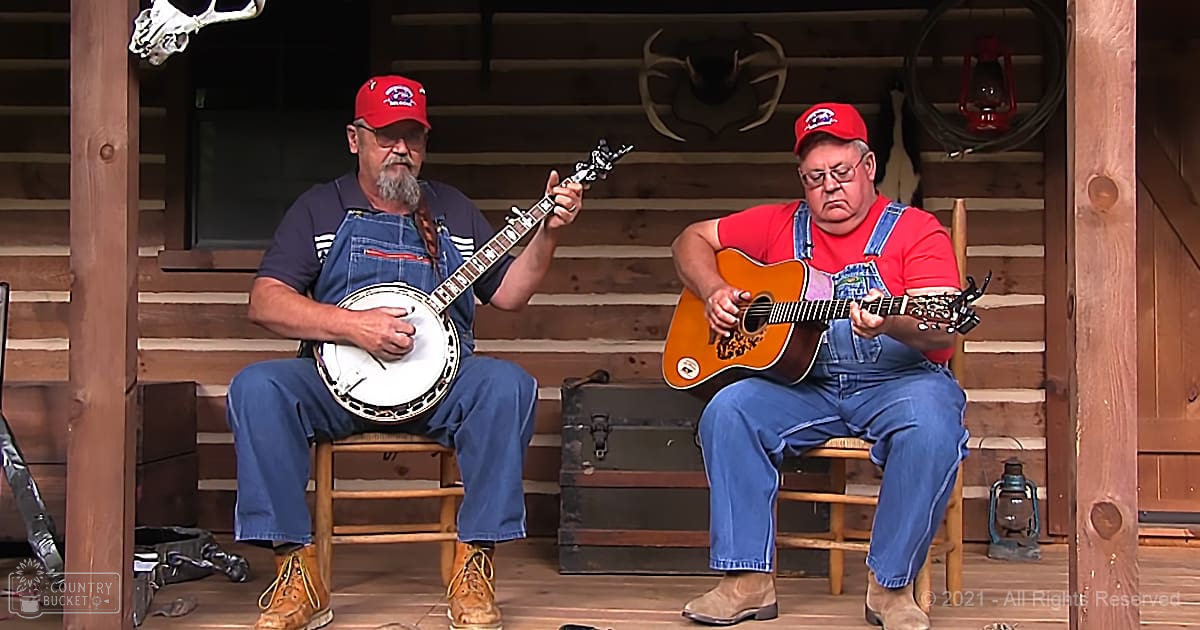 Two middle-aged to older adult males playing musical instruments in a rustic cabin porch setting, surrounded by wooden furniture and antler decorations.