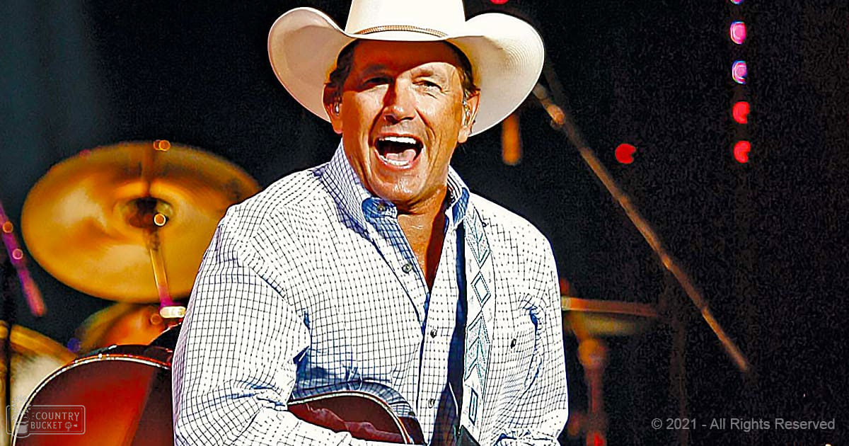 Middle-aged male performer wearing a white cowboy hat, checkered white and blue shirt with denim vest or jacket, singing into microphones in an indoor concert setting with red and purple stage lighting.