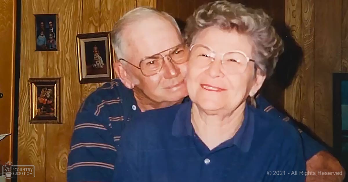 An older couple indoors in a home setting; man wears striped sweater, woman in navy-blue shirt with thin-framed glasses; cozy wooden paneling and framed photos suggest a family room.