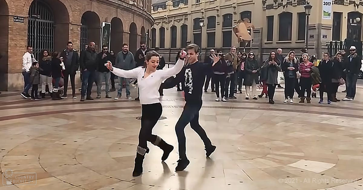 A couple dancing in a public square with historical architecture under soft evening light; people of various ages wearing winter clothing.