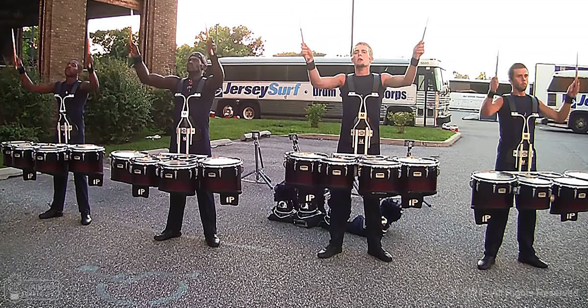 A group of young adult male drummers performing outdoors in front of buses labeled "Jersey Surf Drum Corps." The scene is set on a paved area with greenery and trees, under clear skies.