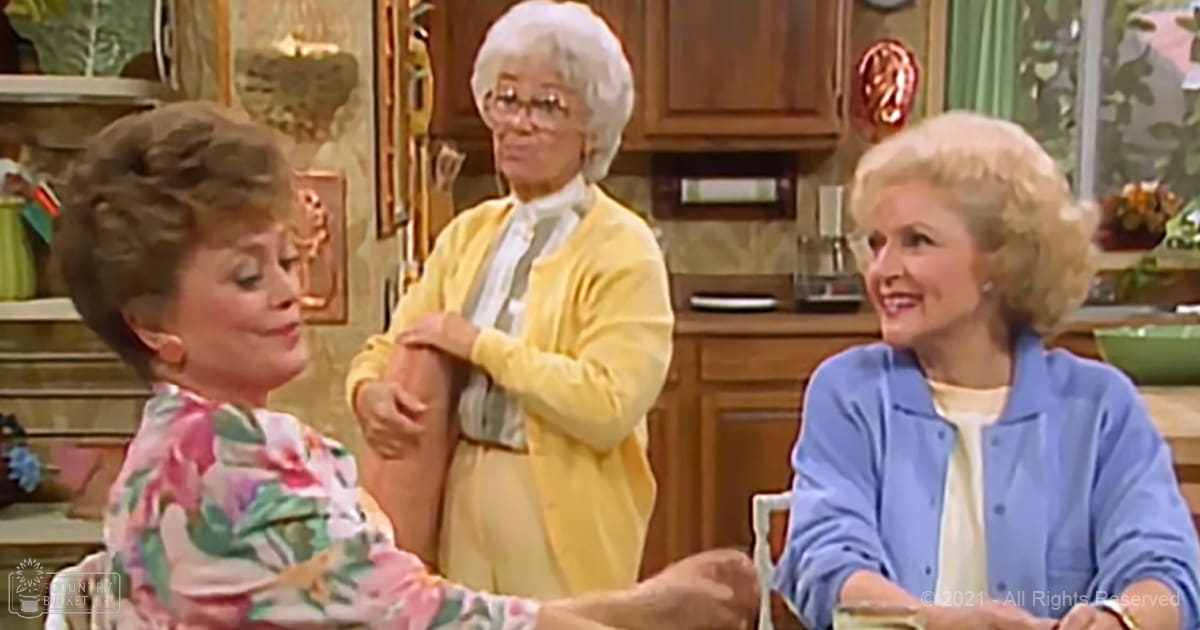 An adult female seated at a table in a home kitchen, wearing a floral-patterned top with short sleeves and holding a small object; another older adult female stands center, dressed in a yellow cardigan over a white blouse with a high collar and tie-like detail; an older adult female sits to the right, in a blue cardigan over a white top with long sleeves and styled hair.