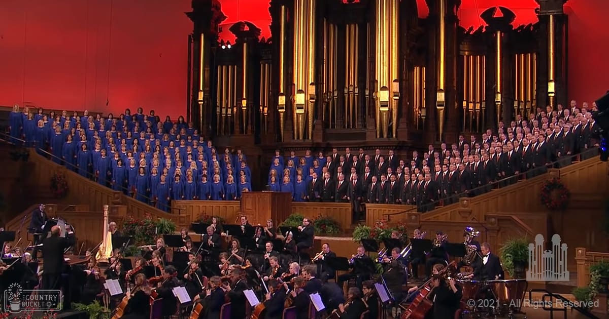 A choir and orchestra performing in a grand hall with red lighting, festive wreaths on the walls, and wooden risers for seating.