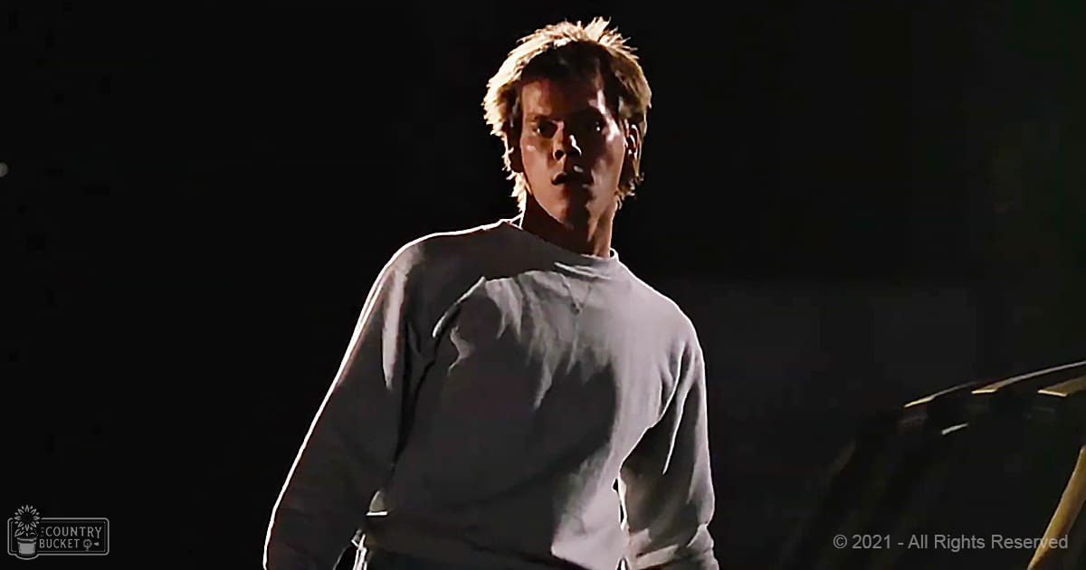 Young adult male in a relaxed white long-sleeved shirt with rolled-up cuffs, standing indoors under dramatic stage lighting.