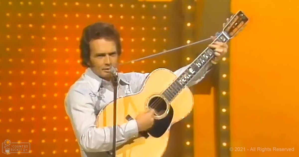 Adult male singer wearing a white long-sleeved shirt with patterned design and tie performing on stage holding an acoustic guitar under orange lighting.