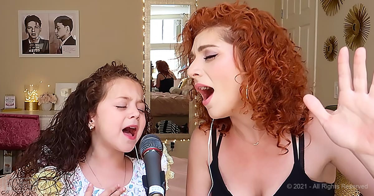 A young girl sings into a microphone with an adult woman in a cozy bedroom setting with string lights and home decor.