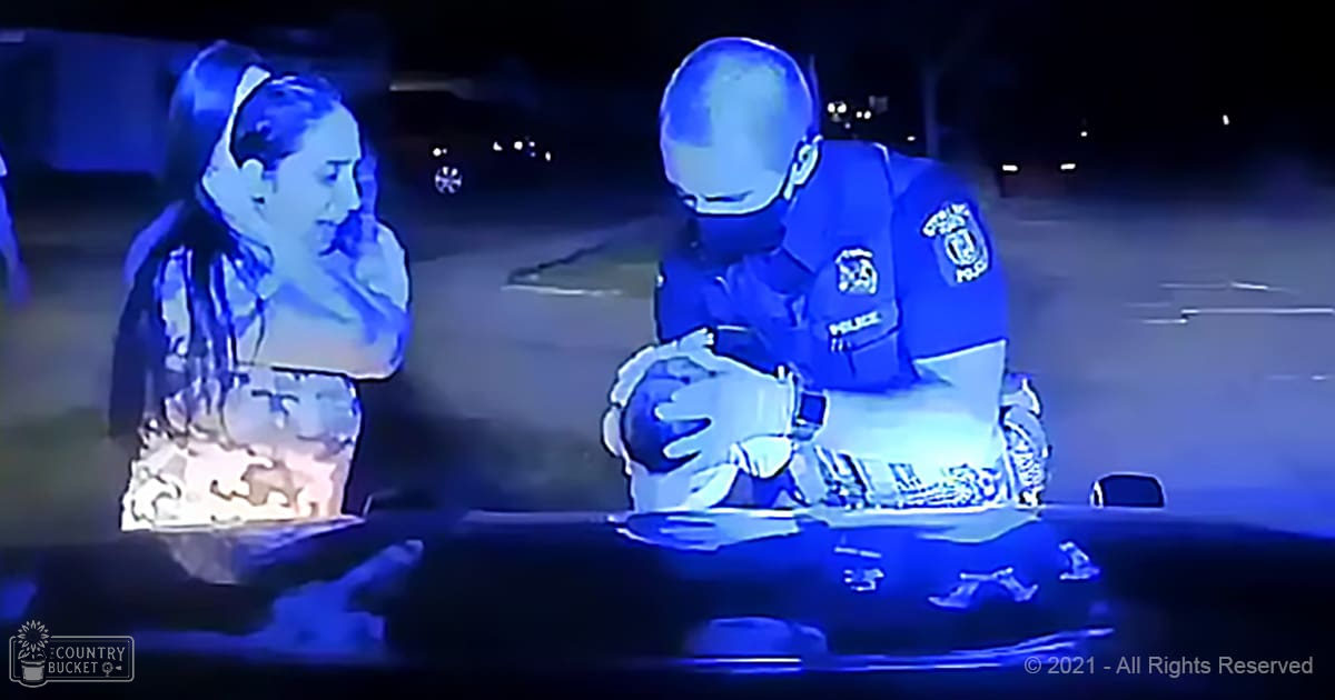An adult male police officer in a dark blue uniform interacting with a young adult female in a light-colored top, seated inside a vehicle at night in an outdoor setting.