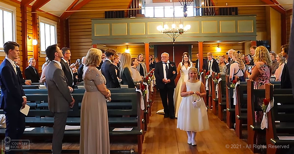 A young girl in a white floral-patterned dress walks down an aisle at a wedding ceremony amidst seated adults and church decor.