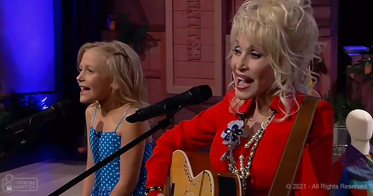 A middle-aged woman singing alongside a young girl in an indoor studio setting, wearing a red shirt with floral brooch and pearl necklace.