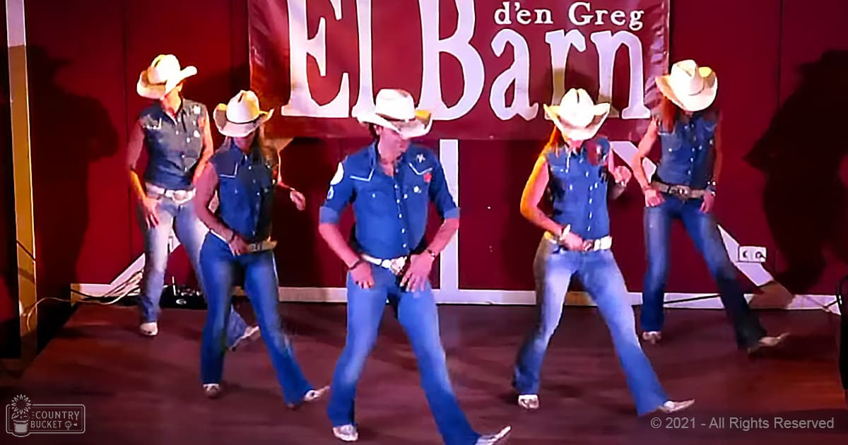 Group dance performance at 'El Barn' with individuals wearing denim vests, blue jeans, white cowboy hats, and belts in a Western-themed setting indoors on a stage or dance floor.
