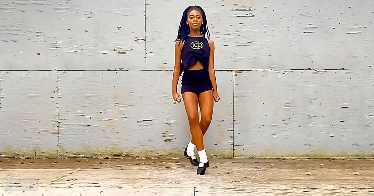 A young adult female in a modern, form-fitting two-piece outfit stands outdoors against a weathered wall during late afternoon or early evening light.