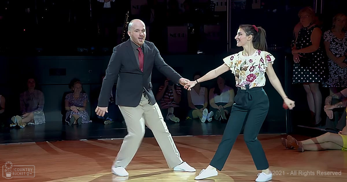 Two dancers in a dance studio, man wearing dark blazer and red tie, woman with floral top and ruffled sleeves performing together under overhead lights.