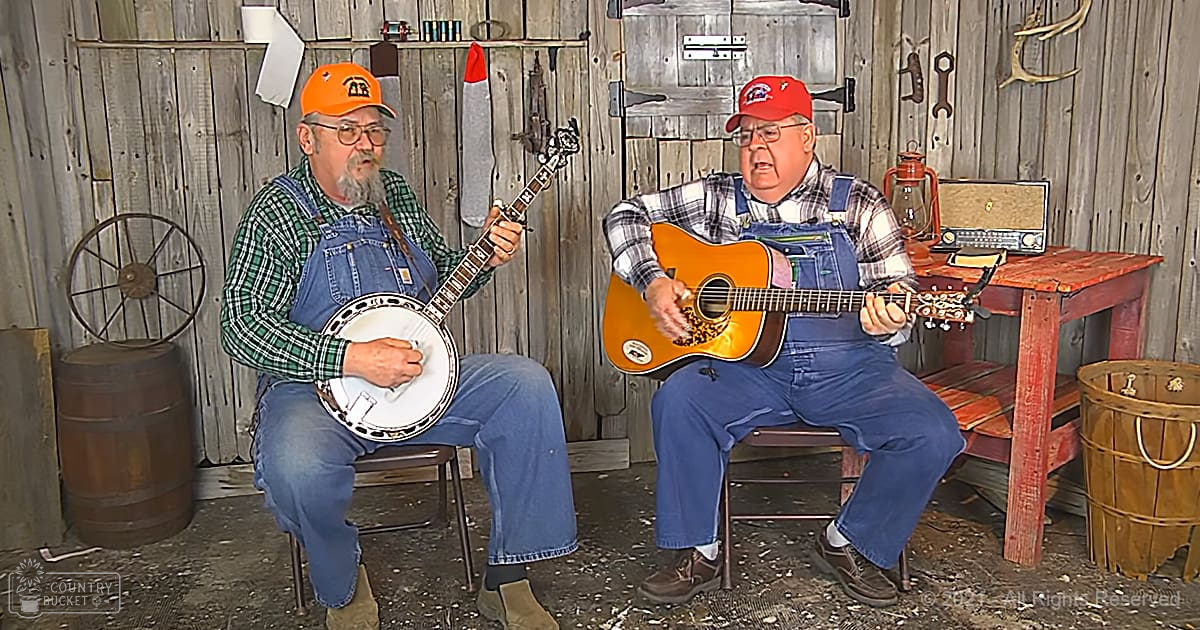 Two older adults play musical instruments in a rustic indoor cabin setting with wooden furniture, tools, and natural light.