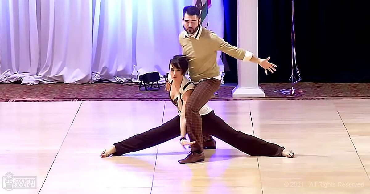 Dance duo performing on a stage with light-colored flooring, white curtains as backdrop, and part of a column suggesting an indoor venue like a ballroom or event hall.