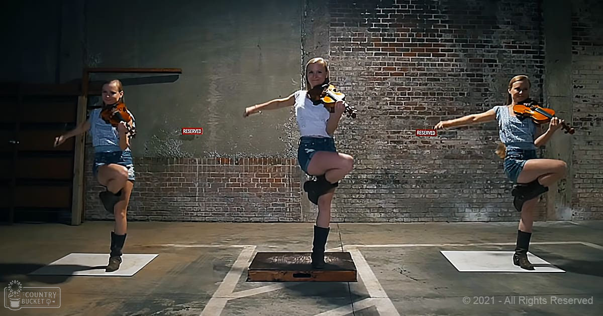 Three young adult females playing violins in an urban setting with a brick wall backdrop and 'RESERVED' signs, natural daylight illuminating the scene.