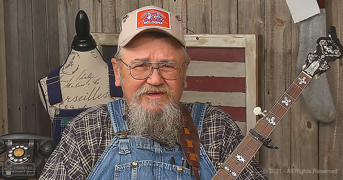 An older adult male in a rustic indoor setting, wearing a plaid shirt with denim overalls and a beige baseball cap, surrounded by vintage decor including a rotary phone and an American flag-themed sign.