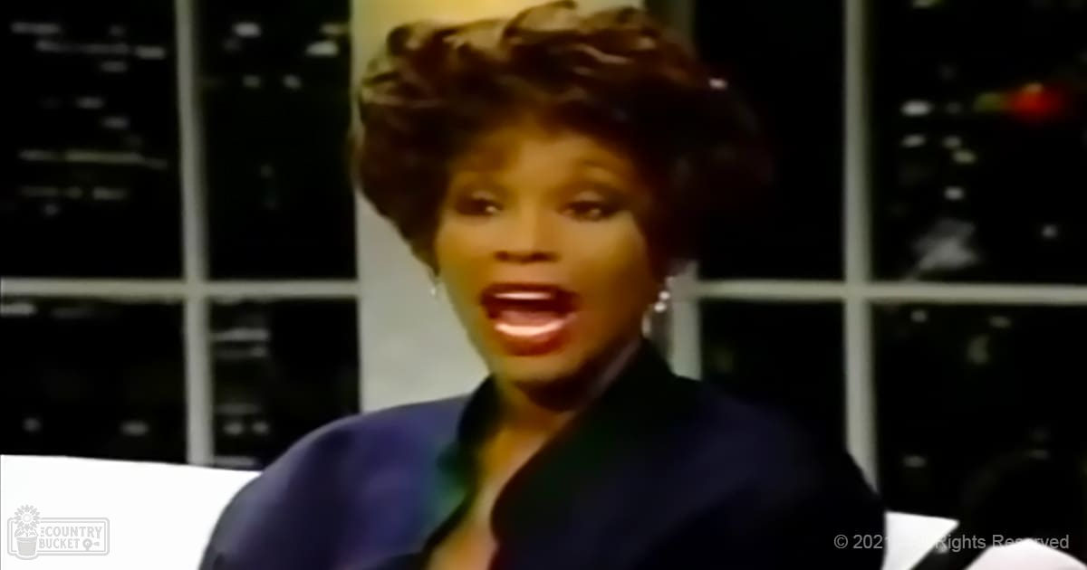 An adult female in an indoor setting, wearing a dark blue blazer with a subtle gradient to purple at the collar and long-sleeved sleeves. She is accessorized with earrings.