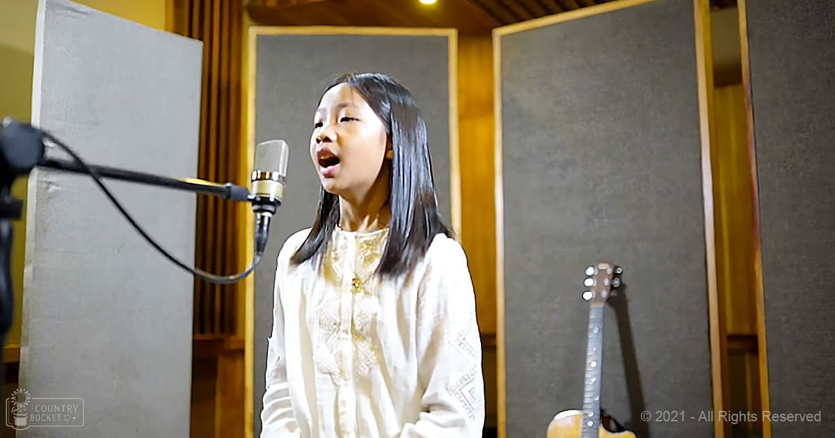 A young female child in a white blouse with lace detailing, standing in a recording studio surrounded by acoustic foam and musical equipment.