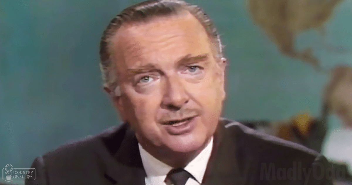 An older adult male in a formal setting, wearing a mid-20th century dark suit jacket with white shirt and tie, standing indoors possibly in an office or studio environment.