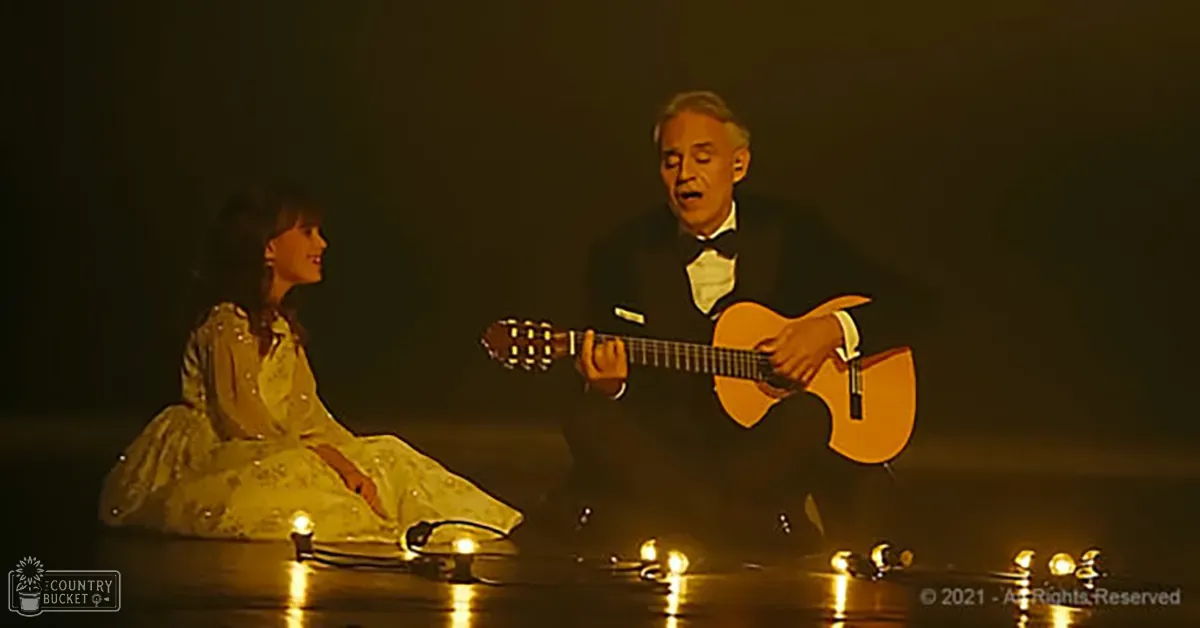 A musical performance scene with two individuals on stage; one in a traditional embroidered dress and another in a formal tuxedo, surrounded by candles creating a warm ambiance.