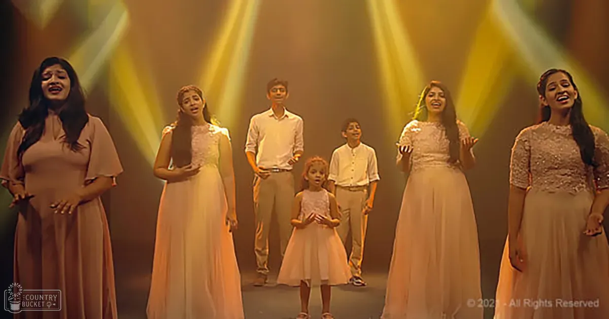A group of adults and children in a studio or theater setting with warm lighting and dramatic yellow beams, wearing light pink dresses and white shirts with beige pants.