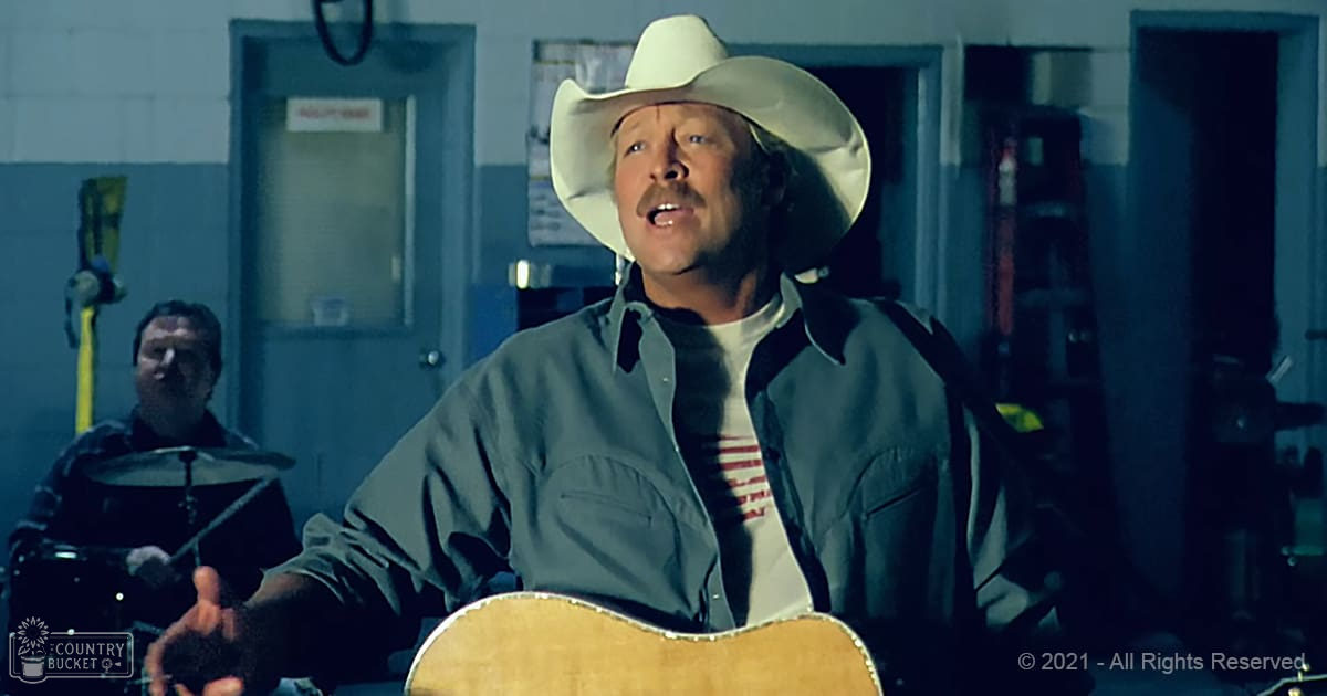 A middle-aged man in a light-colored cowboy hat plays guitar indoors with tools visible in the background.
