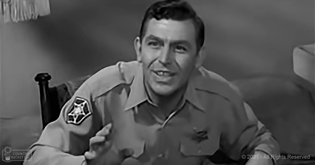 A middle-aged adult male wearing a light brown/tan uniform shirt with patches, standing indoors in an environment with minimal decor and neutral-colored walls. The setting suggests evening or night due to the lack of bright daylight.