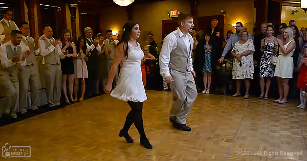 A couple dancing at an indoor wedding reception with a mix of adults in formal attire, warm lighting, and decorative columns.