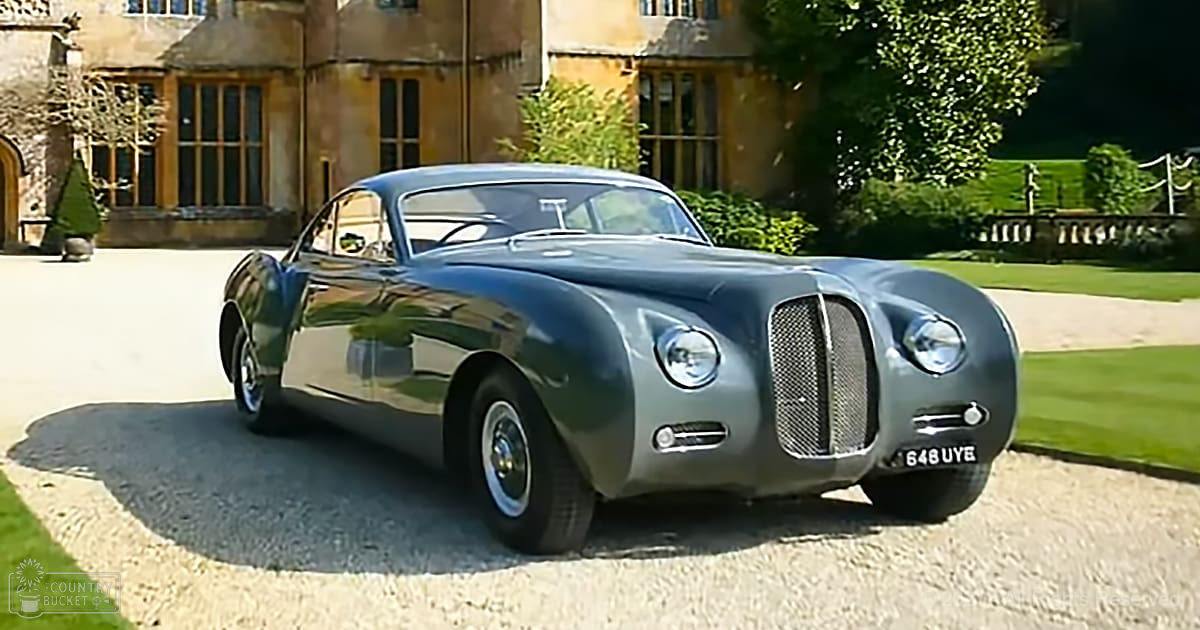 A vintage car with rounded body and large headlights is parked in front of an estate or mansion, surrounded by well-maintained grounds and lush vegetation.