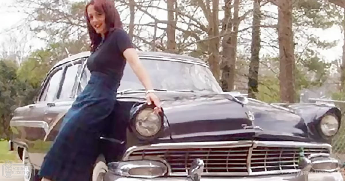 A young adult female in a dark blue dress stands outdoors amidst greenery, with a classic black car as the focal point of the scene.