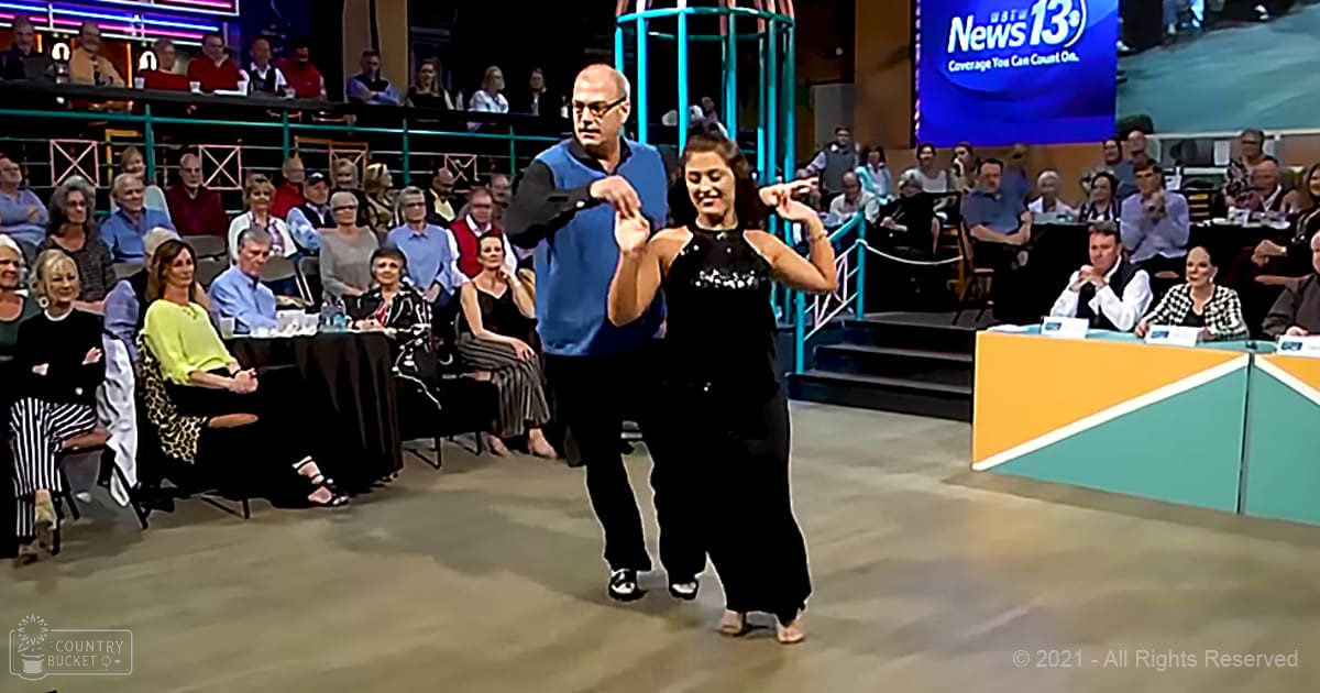 A couple dancing in a studio setting with an audience seated around them, wearing formal attire.