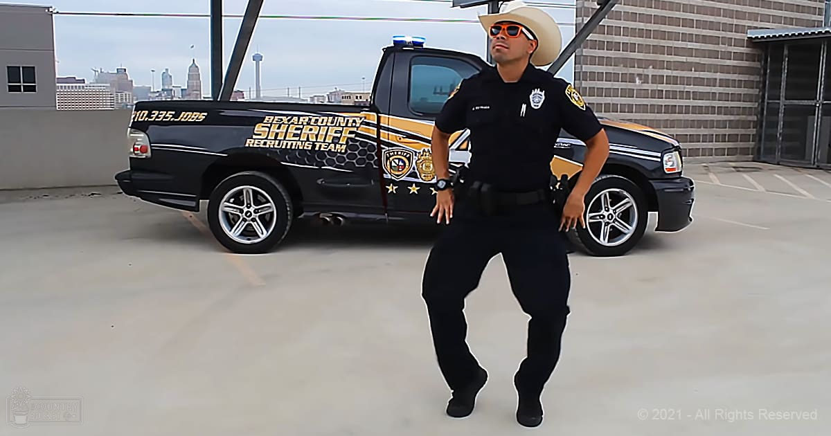Adult male in black police uniform with patches and badges stands before a Bexar County Sheriff's Department truck, wearing white cowboy hat and orange-tinted sunglasses under an overcast urban setting.
