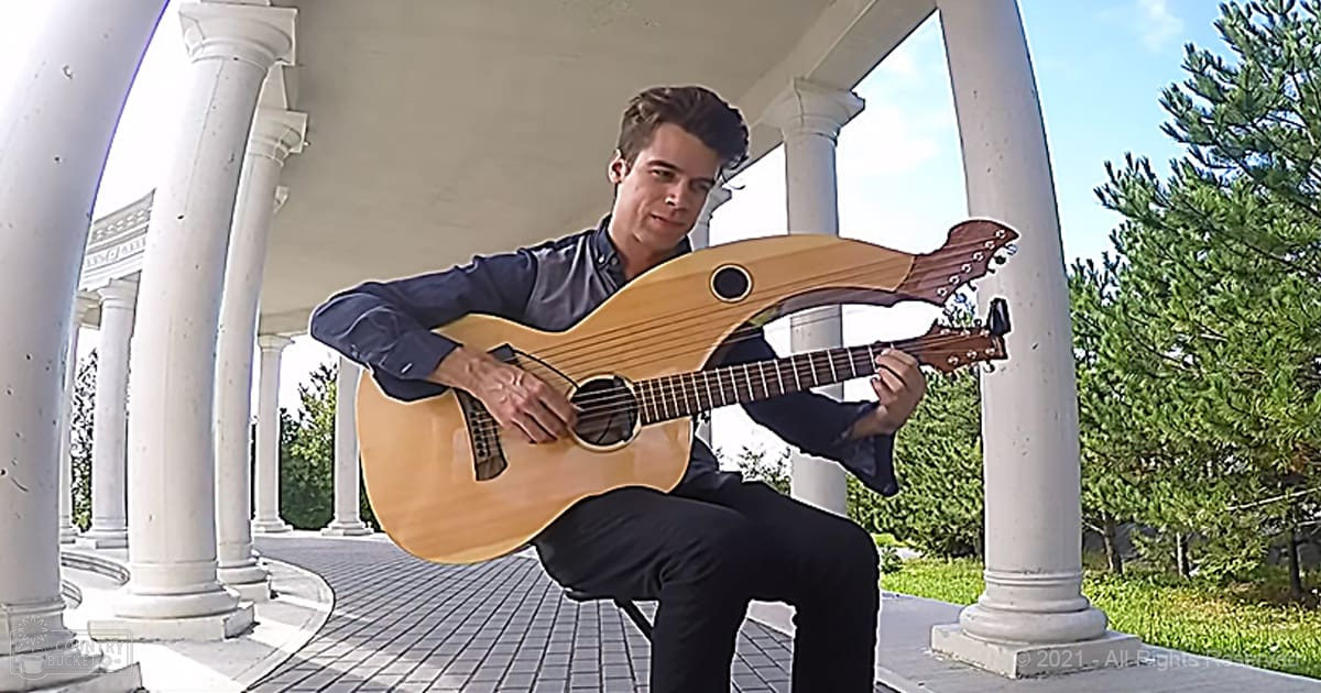 A young adult male playing an acoustic guitar in a classical-style colonnade setting with white columns and greenery, under clear blue skies.