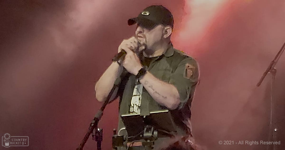 Adult male singer in a dark green military-style shirt with black tactical gear, performing on stage at a concert amidst red smoke effects.