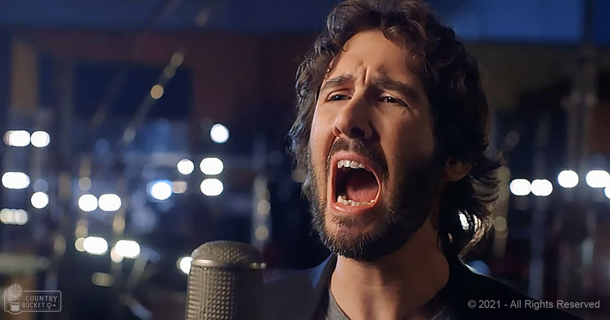 A male adult wearing a dark-colored suit jacket, singing into a microphone in a recording studio at night with artificial lighting.