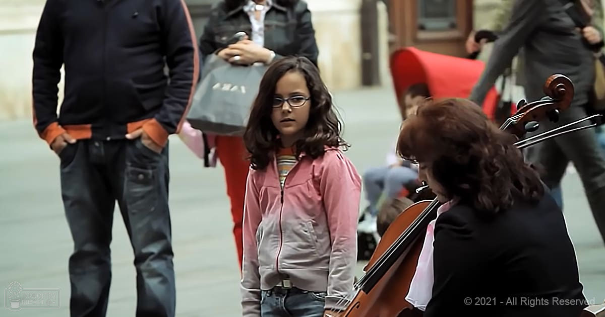 A young girl in a pink zip-up jacket and blue jeans stands outdoors amidst adults and musical activity.
