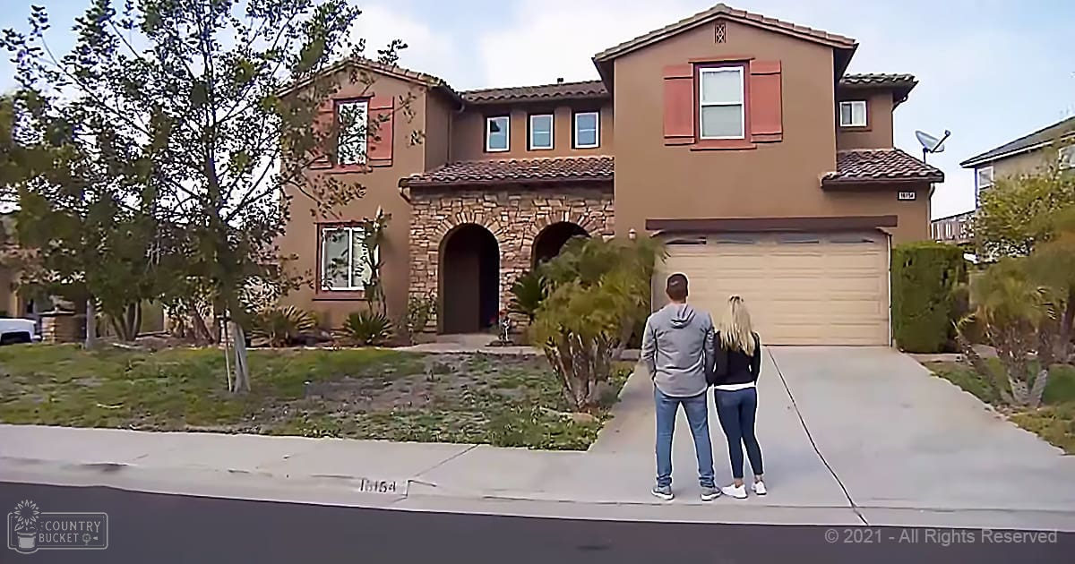 A young adult male singer wearing a gray hoodie and dark blue jeans stands in front of a suburban house with beige walls and red shutters on windows, likely spring or summer given the attire and greenery. The setting includes landscaping with small trees and shrubs.