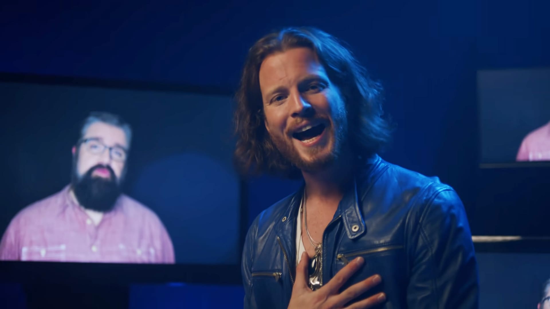 A young adult male with long hair wears a dark blue leather jacket over a white shirt indoors, possibly related to entertainment or media production.