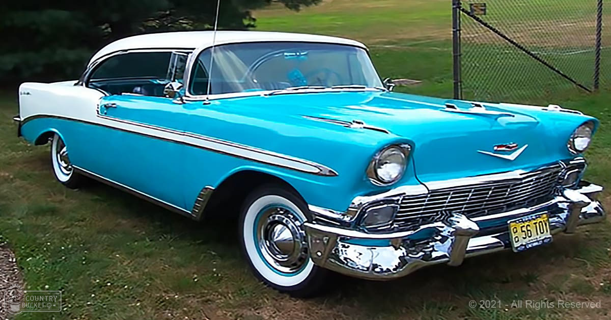 A classic 1950s blue Chevrolet Bel Air with white wall tires, chrome detailing, and a license plate reading "856 T01" parked on green grass outdoors. A chain-link fence and lush trees are visible in the background under natural daylight.