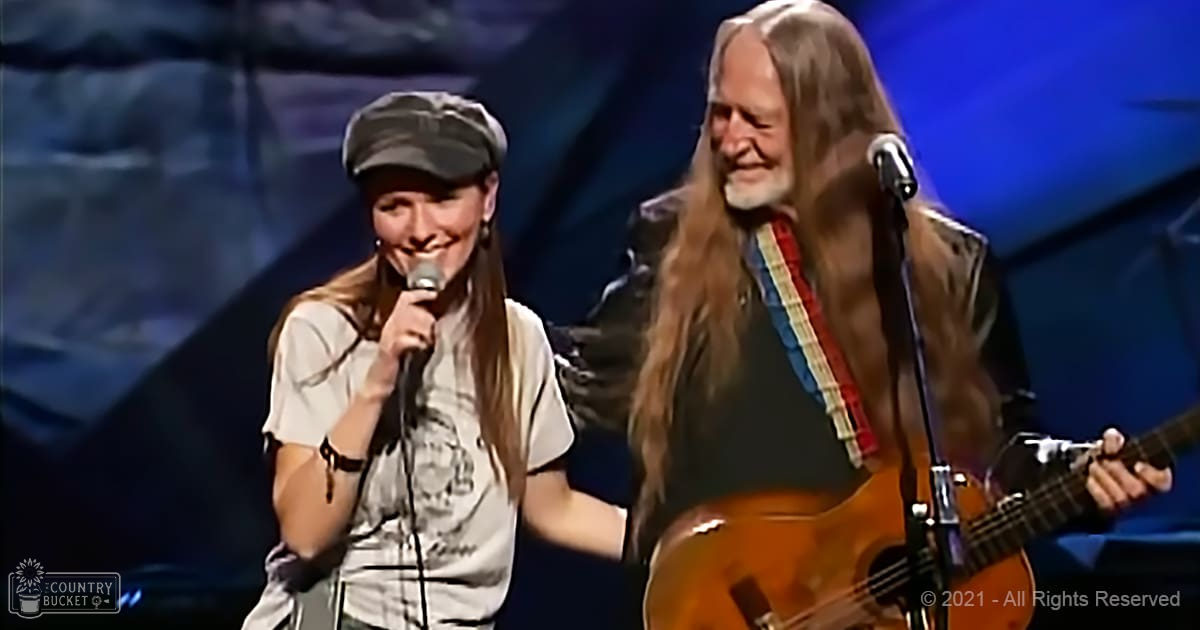 A musical performance scene with a young adult female in a white graphic tee and dark pants, wearing a black backward cap with long hair down her shoulders adorned by a bracelet or wristband on one arm. An older adult male, possibly an adult singer, is seen to the right, dressed in a dark t-shirt with a guitar strap over his shoulder, long hair down to his waist, beard, and potentially a jacket or vest, playing a red and white striped guitar.