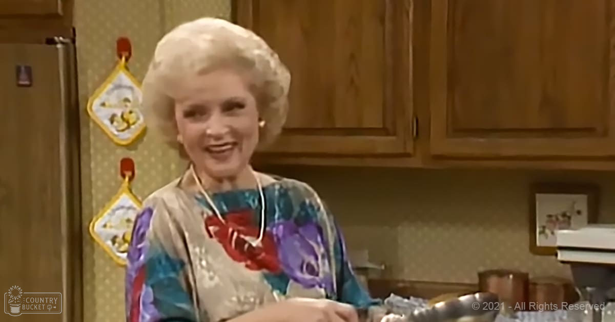 A middle-aged woman wearing a long-sleeved, blue floral blouse with red and purple flowers, standing in a kitchen setting with wooden cabinets and home decor items.