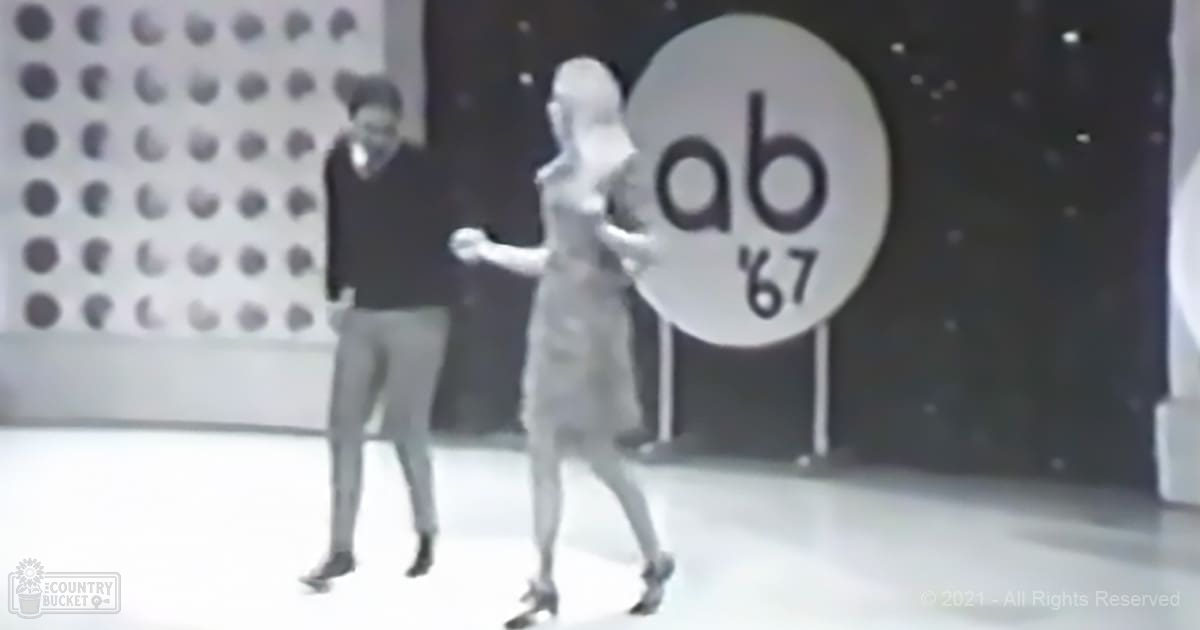 An adult male singer in a dark-colored sweater and light-colored pants with dark shoes stands beside another adult female wearing a knee-length, sleeveless dress made of smooth fabric, possibly silk or satin, with high-heeled shoes. The setting appears to be an indoor studio from 1967.