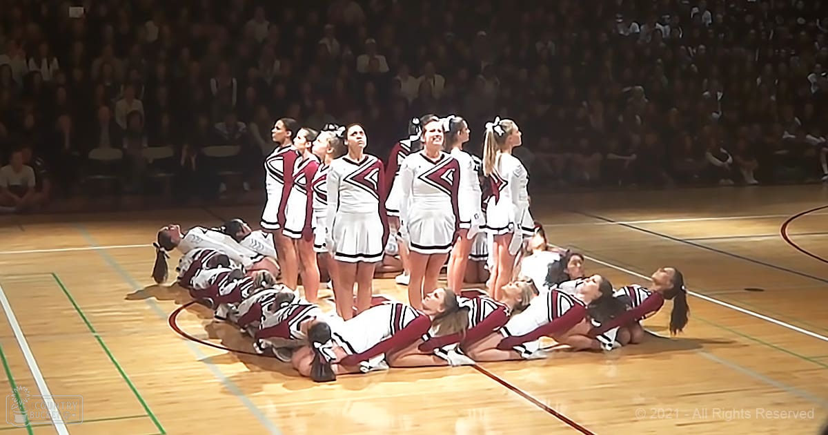 A cheerleading team performing at an indoor sports arena, surrounded by a seated crowd in artificial stage lighting.