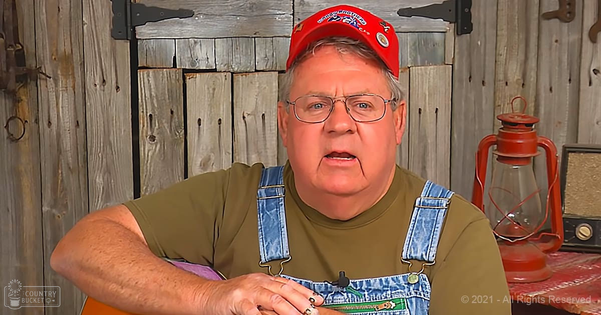 An older adult male in a rustic outdoor setting, wearing an olive green t-shirt with blue denim overalls and holding a guitar strap.