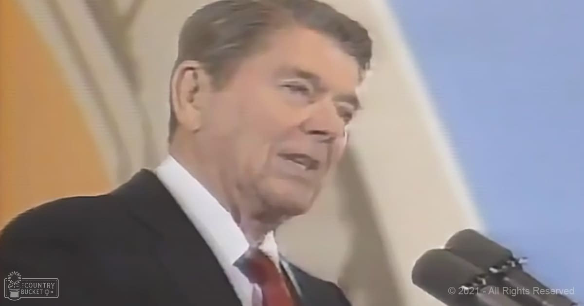 A middle-aged adult male in a formal dark suit jacket with a white dress shirt and red tie speaks at an event, possibly indoors with soft lighting.